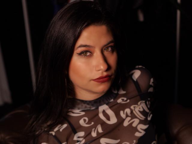 MarianaConti with dark hair, red lipstick, and green eyes, wearing a sheer top with white text, sitting in dim light. Her tits are slightly visible through the fabric.