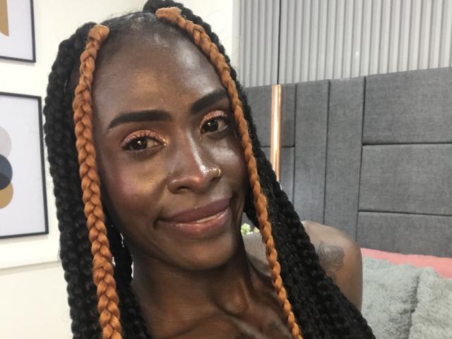 SamaraaJackson69 with dark skin, orange and black braided hair, small nose ring, smiling, makeup, tattoo on right shoulder, gray headboard in background, looking at camera.