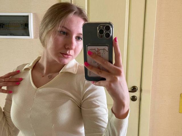 SweetLisaa with pink nails taking a selfie, wearing a beige ribbed shirt, showing her tits, holding a phone with a cute case, blonde hair, light skin, and a heart necklace.