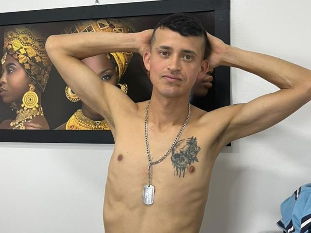 SasusKee, shirtless with a dog tag necklace, arms behind head, showing a chest tattoo, standing in front of a framed picture of dark-skinned women with gold jewelry.