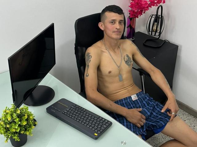 Shirtless SasusKee with tattoos, dog tags, and blue plaid shorts, sitting in a black chair, hand on cock, in front of a glass desk with a keyboard and monitor.