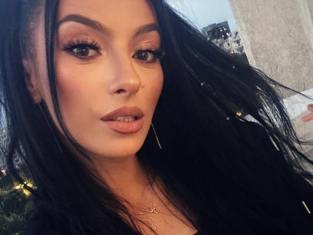 KinkyAmelie with long black hair, brown eyes, full lips, and subtle makeup, wearing gold earrings and a cross necklace, looking slightly to the side. Boob outline visible under black top.