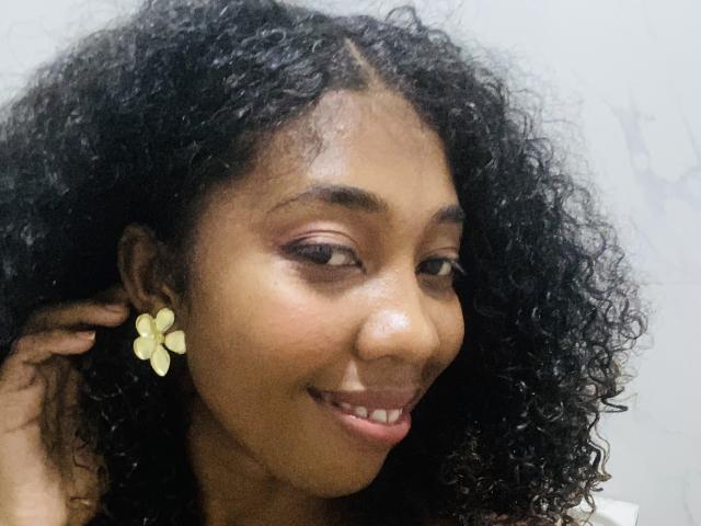 Chore has dark curly hair, brown skin, and is smiling with yellow flower earrings. She's touching her head with one hand, showing off her pink lipstick.