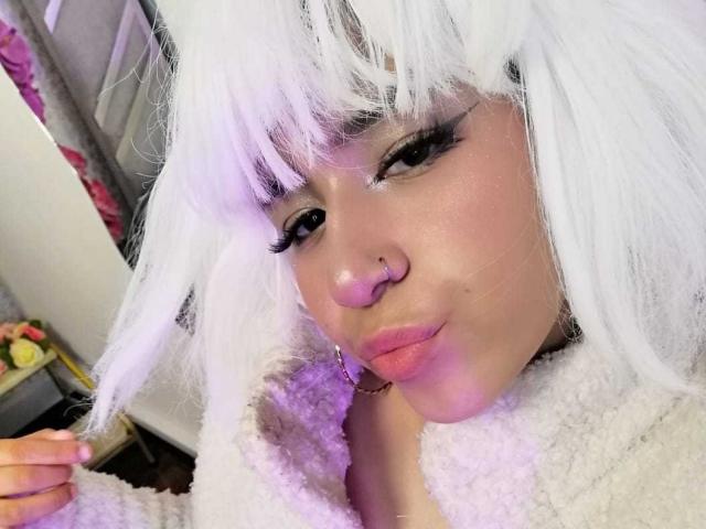 DulceSweett met wit, pluizig haar, dikke zwarte eyeliner, roze lippen, een neusring en een witte, zachte jas, maakt een fronsend gezicht en houdt haar haar vast..