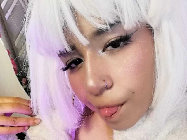DulceSweett has white hair, a nose ring, and is sticking her tongue out with pink lips. Her eyes have thick black eyeliner and long lashes. She's touching her hair.