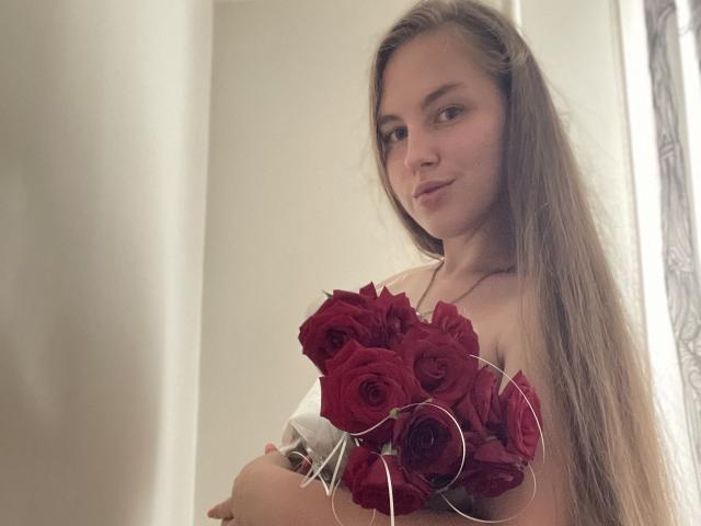 Dooris with long brown hair, holding a bouquet of red roses, smiling slightly, wearing a white off-shoulder top, looking at the camera.