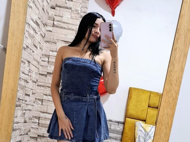 LizzeJoy with black hair, wearing a strapless denim dress, taking a mirror selfie with a pink phone, showing her small tits and tattoo on her left arm.