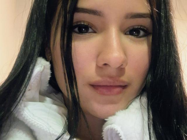 LizzJoy with long black hair, wearing a white fluffy robe, has a cute, slightly pouty face and full lips. Her dark eyes and subtle makeup are noticeable.