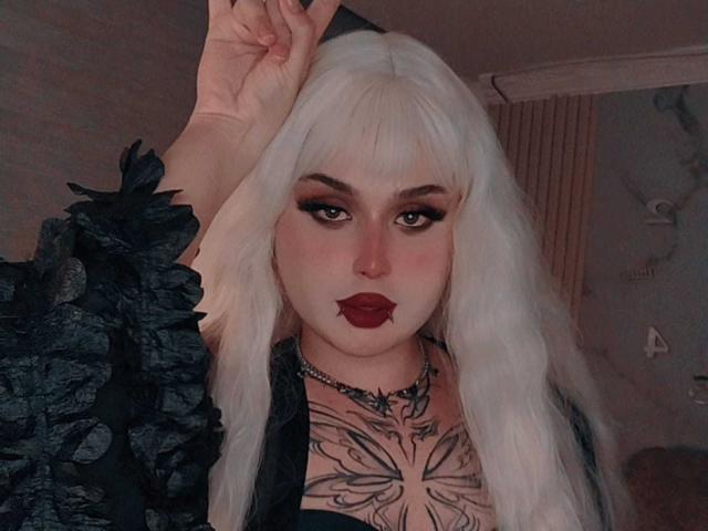 MadameRaven with white hair, dark makeup, and red lipstick, wearing a black ruffled top, exposing a butterfly tattoo on her chest, raising her hand.