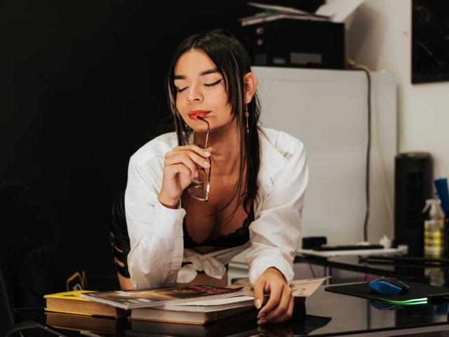 BabeCaty with long black hair, wearing a white shirt, black bra, and glasses, leaning on a desk, touching her red lips, with books and a mouse in the foreground.