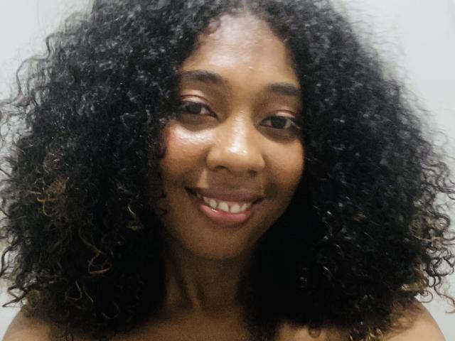 Chore has dark curly hair, brown eyes, and a smile showing white teeth. Her tan skin is visible from the shoulders up. She looks cute and natural.