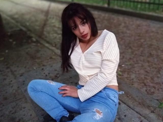 RouseParker with dark brown hair, white striped top, and blue ripped jeans, squatting on a concrete path, looking at the camera with a slight pout.