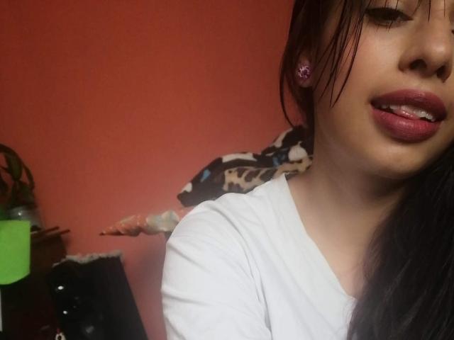 RouseParker's face, showing dark lipstick and slightly open mouth, wearing a white shirt, with long black hair and pink earrings, in a red-walled room.