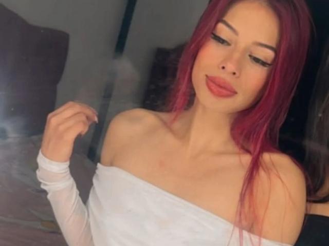 RouseParker with red hair, off-shoulder white top, and eyeliner, looking down with slightly parted lips, showing her small tits and slim collarbone. She's touching her hair.
