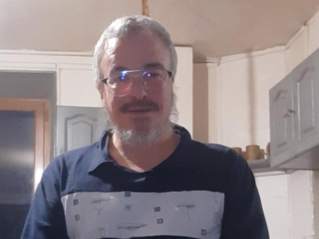 TitiWx has gray hair, a beard, and glasses. He's wearing a black and white shirt with a pattern. He's standing in a kitchen. His ass is not visible.