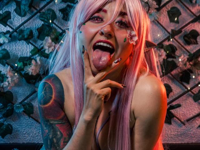 MeiJaisen with pink hair, elf ears, and tattoos, sticking out tongue, finger on cheek, red and blue lighting, flower in hair, and colorful face paint. Her tits are visible.