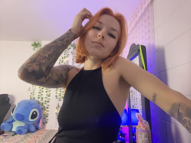 SarahRousen with red hair, tattooed arm, black halter dress, showing her tits, looking at camera, in bedroom with blue Kigyo plushie.