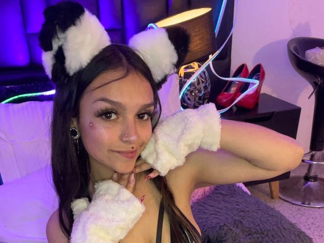MarietteJones with white furry cat ears, wrist cuffs, black top, long brown hair, cute face, nose piercings, and a confident smile. Red heels and black stool in background.