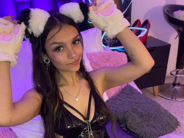 MarietteJones with dark hair, wearing black shiny top, cat ears, and fluffy pink-and-black paw gloves, raising arms, showing small tits, nose ring, and cute expression, in a purple-lit room.