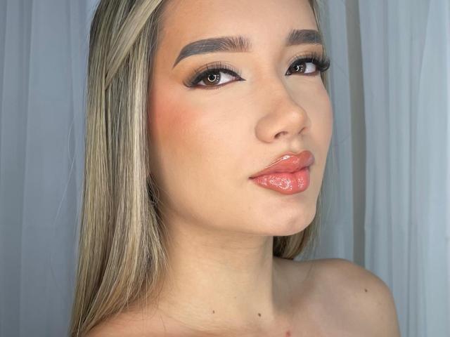 LuSorel has shiny blonde hair, thick eyebrows, and glossy pink lips. Her brown eyes are framed by long, dark eyelashes. She's giving a slight pout.