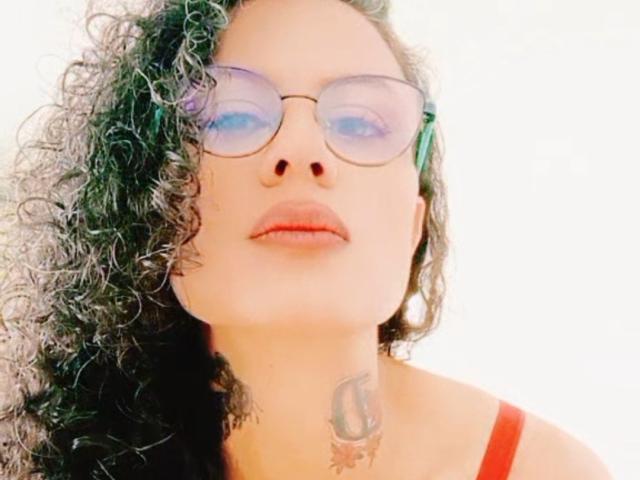RosannaBlue with curly green and black hair, wearing purple-tinted glasses, red lipstick, and a red top, showing a tattoo on her neck, looking up with a slight smile.