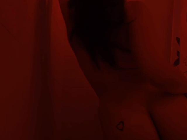 AshleyDaphna, con il petto scoperto e un piccolo tatuaggio a forma di cuore vicino al seno, illuminata da luci rosse, mostra il profilo laterale con i capelli scuri che coprono parzialmente il viso. I capezzoli sono leggermente visibili..