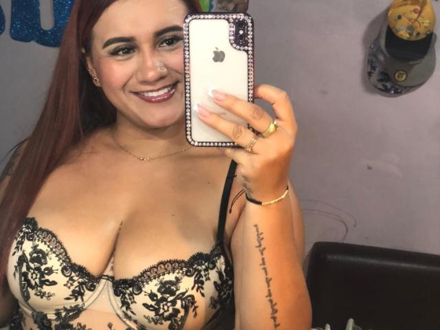 AmberDesire mit großen Brüsten in einem Spitzen-BH, lächelnd und ein Selfie mit einem iPhone machend. Sie hat langes, rotes Haar, ein Tattoo am rechten Arm und trägt einen Ring..