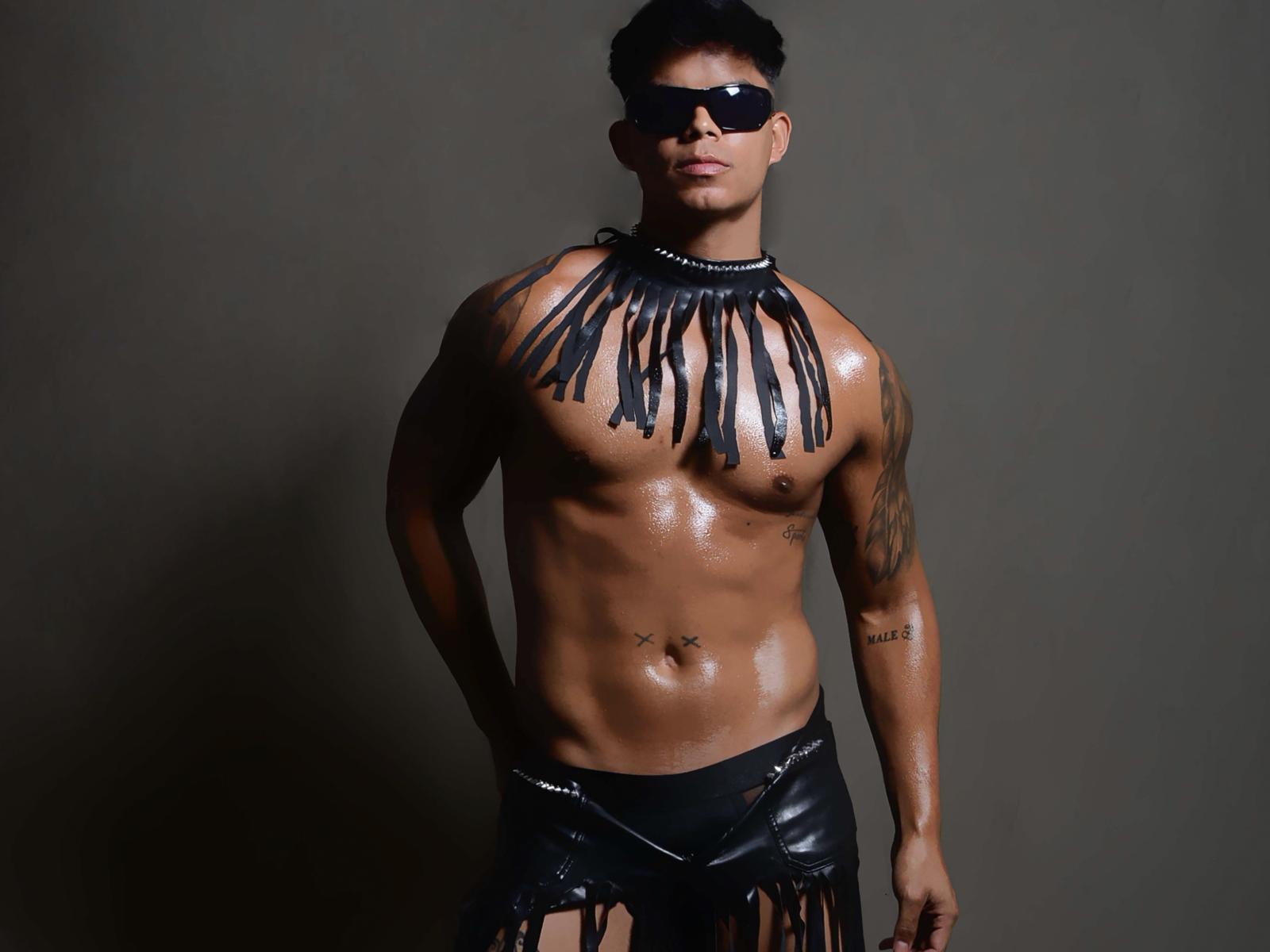 Shiny, muscular XerosCl with black leather chest fringes, black leather skirt, shiny oil-covered skin, tattoos, black sunglasses, and short black hair, standing against a dark background.