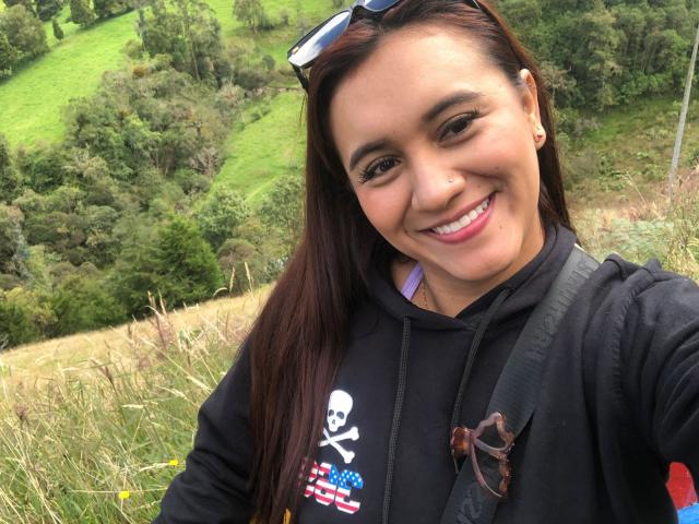 AmberDesire, mit einem Lächeln und pinkfarbener Lippenstift, hat dunkelbraunes Haar, trägt eine schwarze Kapuzenjacke mit einem Totenkopf- und Flaggenmuster und hat einen Nasenring. Ihr Hintern ist nicht sichtbar..