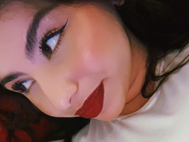 Il viso di VenusxMusa, con sopracciglia scure, lunghe ciglia, rossetto rosso e un leggero sorriso, mostra capelli castano scuro parzialmente visibili. La sua pelle è chiara con un leggero rossore..