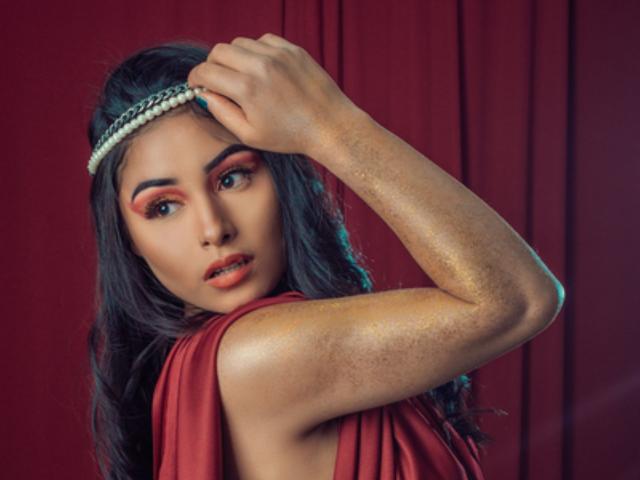 EmilyVv has shiny gold arms, dark wavy hair, red makeup, and a pearl headband, looking over her shoulder with a slightly open mouth. Red background.