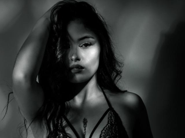 EmilyVv with long, messy hair, wearing a lace bra, has her arm raised, shadowing one eye, and showing off her tits and a small tattoo on her chest. Black-and-white.