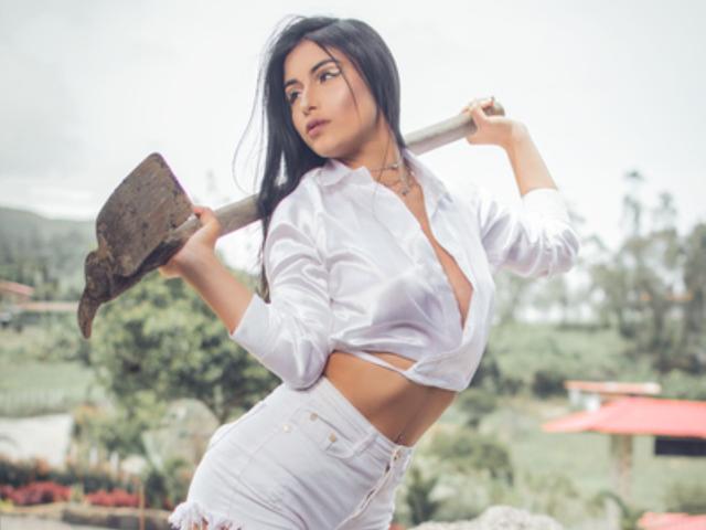 EmilyVv with long black hair, white button-up shirt revealing her tits, holding a dirty axe on her shoulder, wearing white jeans, standing outdoors, looking to the side.