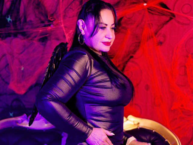 LucianaBlack with black wings, wearing a tight black mesh top, showing her big tits, red lipstick, and hoop earrings, posing confidently with a slight smile.