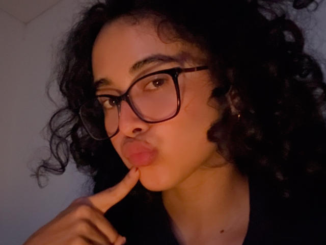 VivienneLaurent with curly black hair, wearing glasses, puckering her lips, touching her bottom lip with her finger, looking at the camera. She has a slight tan and dark eyebrows.