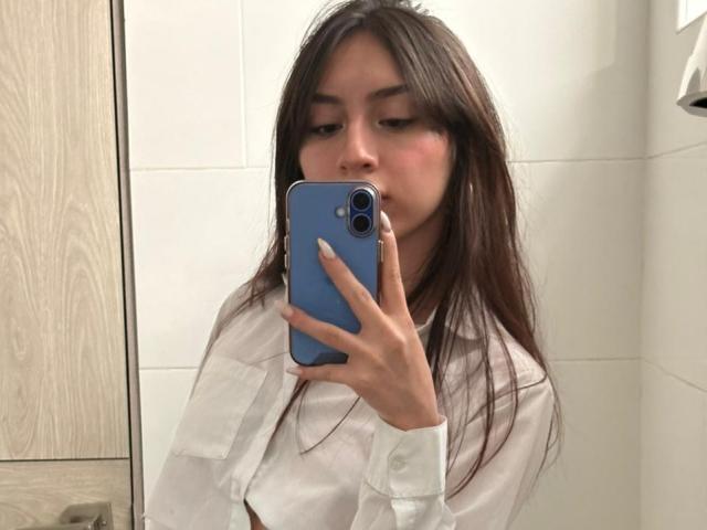 Luluberrie with long brown hair, white shirt, holding blue phone, taking selfie in mirror, showing slight blush on cheeks, looking down, in bathroom.