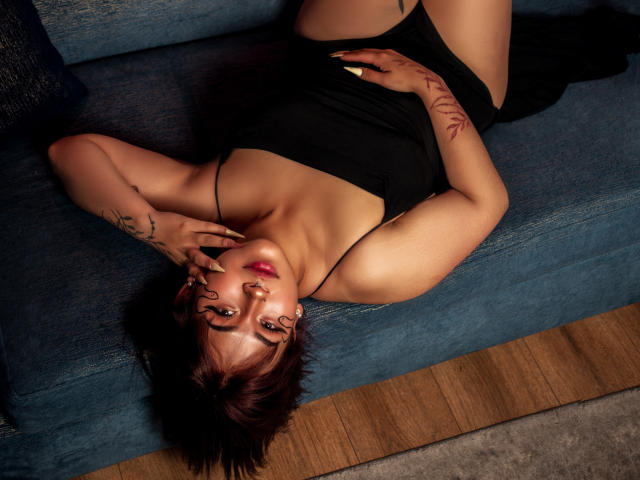 KionI with short brown hair, lying on a blue sofa, wearing a black dress, showing tattoos, holding her face, and looking at the camera, showing her tits.