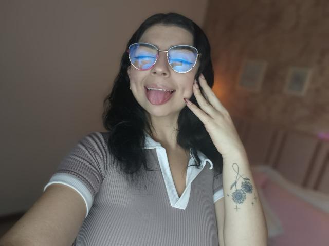 AnntonellaWatson with black hair, wearing glasses reflecting blue light, sticking out tongue, hand near face, tattoo on right wrist, in ribbed brown shirt with white trim.
