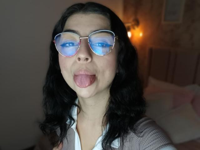 AnntonellaWatson with blue-eyed glasses, black wavy hair, and tongue out, wearing a white shirt and gray cardigan, in a dimly lit room.
