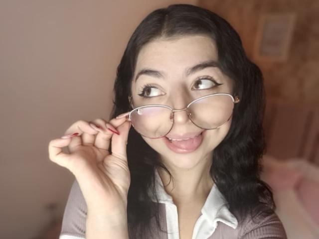 AnntonellaWatson with black wavy hair, wearing glasses, sticking out tongue, and holding glasses with red nails, smiling, in a pink shirt, showing her cute face.