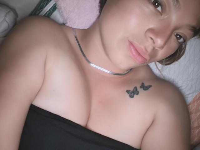 ScarlettAmelia has light skin, brown eyes, and pouty pink lips. She's lying down with black strap covering her tits, showing butterfly tattoos on her right shoulder.