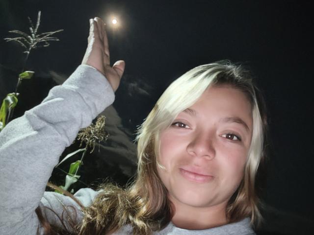 ScarlettAmelia with blonde and brown hair, wearing a grey long-sleeve shirt, raising her right hand to a bright light in a dark night setting, smiling slightly.