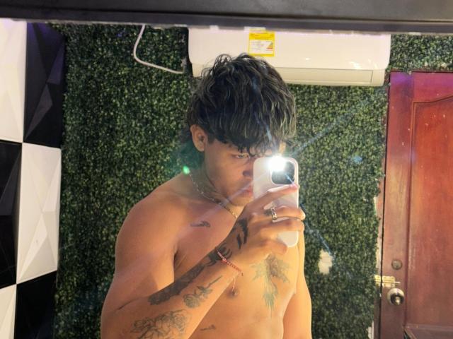 Dave1xAbby sans chemise, avec des tatouages, tenant un téléphone, autoportrait dans un miroir, cheveux noirs ondulés, alliance à la main gauche, mur texturé vert, porte en bois, unité de climatisation blanche au-dessus..
