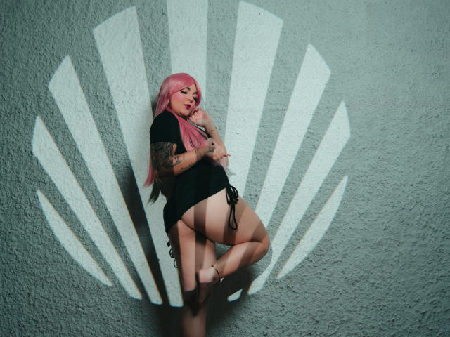 MelodyBlossom with pink hair, black shirt, and tan thong, tattooed arm, standing against white fan-shaped wall, legs bent, looking at phone, bare-ass visible, pink lipstick, medium tits.
