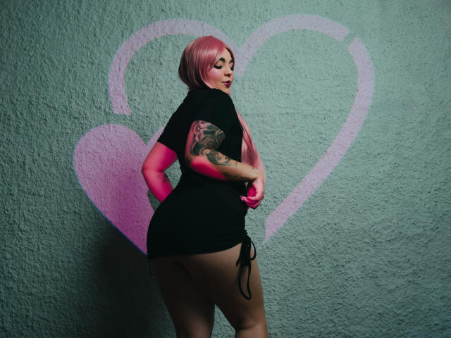 MelodyBlossom, avec ses cheveux roses, sa robe noire et ses tatouages, montrant ses formes généreuses, debout, les mains sur les hanches, regardant en arrière avec un léger sourire, un graffiti mural en forme de cœur rose..