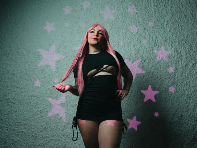 MelodyBlossom, con cabello rosa, vestida con un vestido negro con aberturas, de pie con las manos en las caderas, mostrando sus pechos, frente a una pared verde con grafitis de estrellas rosas..