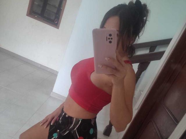 CharlotteDilucca with dark hair in a messy bun, wearing a red sports bra and black shorts with green polka dots, taking a selfie with her pink iPhone.