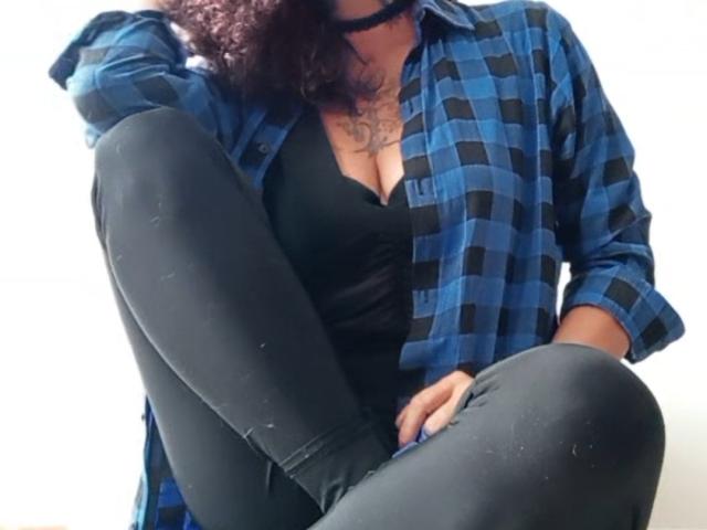 RosannaBlue, met krullend bruin haar, draagt een blauw geruit shirt, een zwarte choker, een zwart tanktopje en strakke zwarte broek, zittend met één knie opgetrokken, waarbij haar borsten zichtbaar zijn..