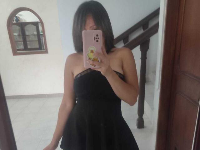 Charlotte Dilucca usando um vestido preto sem alças, tirando uma selfie no espelho com o celular, cobrindo os seios, mostrando o bumbum no reflexo, com cabelo castanho escuro..