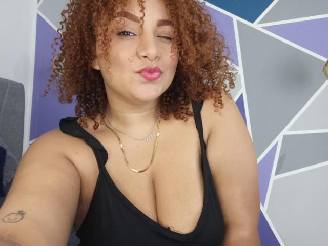 YeraCarey has curly red hair, puffy pink lips, a gold necklace, and a black tank top showing her big boobs. She's winking and sticking out her tongue.